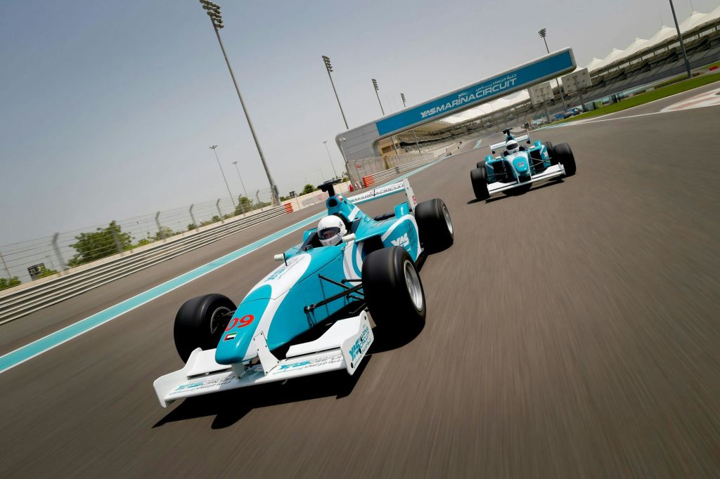 야스 마리나 서킷 (Yas Marina Circuit) 표지판이 보이는 맑은 하늘 아래 레이스 트랙에서 파란색과 흰색의 포뮬러 레이스 카 두 대가 과속하고 있습니다.