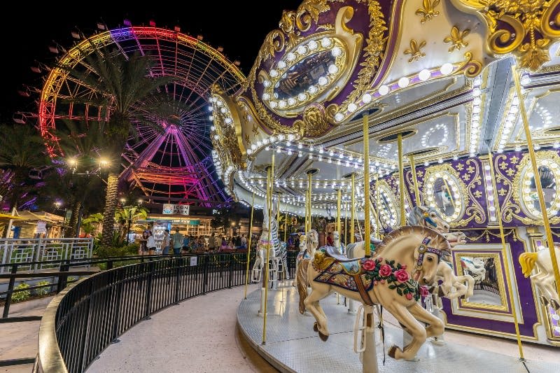 밤에는 밝은 회전목마와 대관람차, 전경에는 화려한 말들이, 배경에는 군중이 있습니다.