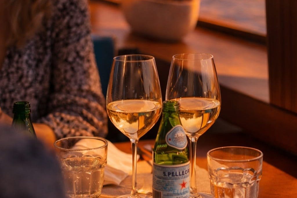 배경이 흐릿한 따뜻한 조명의 테이블에 화이트 와인 두 잔과 스파클링 워터 한 병이 놓여 있습니다.