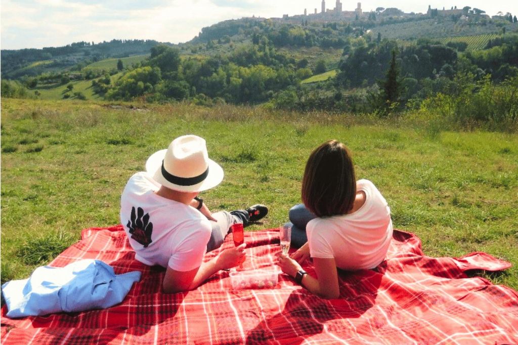 화창한 날 멀리 언덕 꼭대기 마을이 내려다보이는 잔디밭에서 빨간색 피크닉 담요에 앉아 있는 두 사람.
