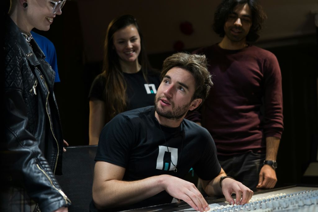 세 사람이 실내에서 교류하고 있으며, 한 사람은 앉아서 고개를 들고 두 사람이 서 있습니다. 캐주얼한 복장, 어두운 배경.