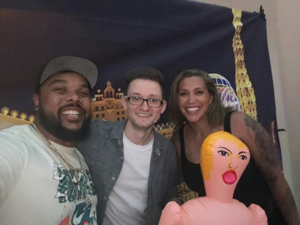 세 사람이 카메라를 보며 미소를 짓고 있으며, 폭발 인형을 앞에 두고 도시 풍경을 배경으로 사진을 찍고 있습니다.