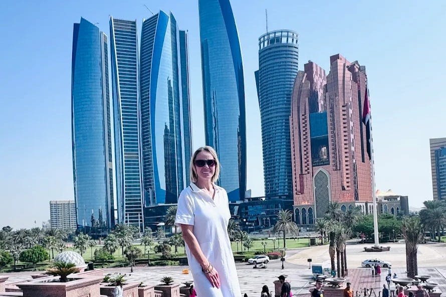 흰색 드레스를 입은 여성이 야외에서 현대적인 고층 건물과 야자수를 배경으로 서 있습니다.