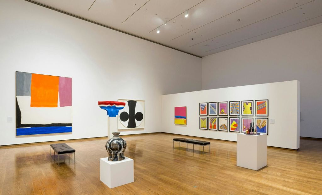 흰색 벽에 추상화, 받침대에 생동감 넘치는 꽃병, 나무 바닥에 벤치가 있는 현대 미술관.