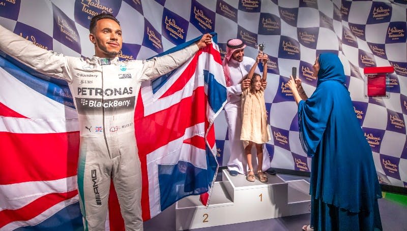 커다란 영국 국기를 들고 있는 흰색 레이싱복을 입은 사람이 "마담 투소" 라고 적힌 배경 앞에 서 있습니다.