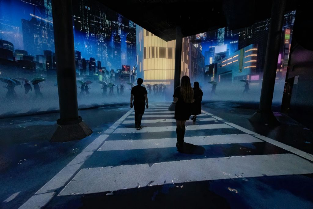 밝은 건물과 디지털 프로젝션이 있는 미래 지향적이고 안개가 자욱한 도시 풍경 속에서 사람들이 얼룩말 횡단을 하고 있습니다.