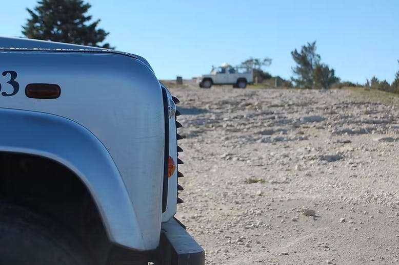 맑고 푸른 하늘 아래 배경에 험준한 지형과 흰색 차량이 있는 은색 차량의 전면 모습.