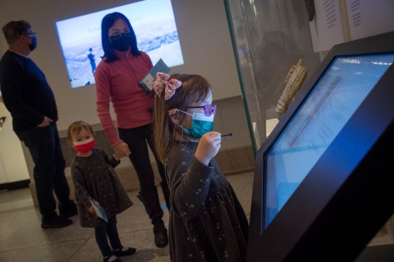 한 어린 소녀가 박물관의 터치스크린 디스플레이와 상호작용하고 있으며, 마스크를 착용한 다른 두 사람이 배경을 관찰하고 있습니다.