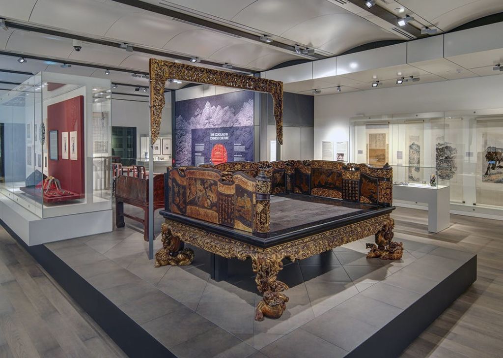 디테일한 조각과 황금색 액센트가 돋보이는 박물관에 화려한 앤티크 침대가 밝은 조명 아래 전시되어 있습니다.