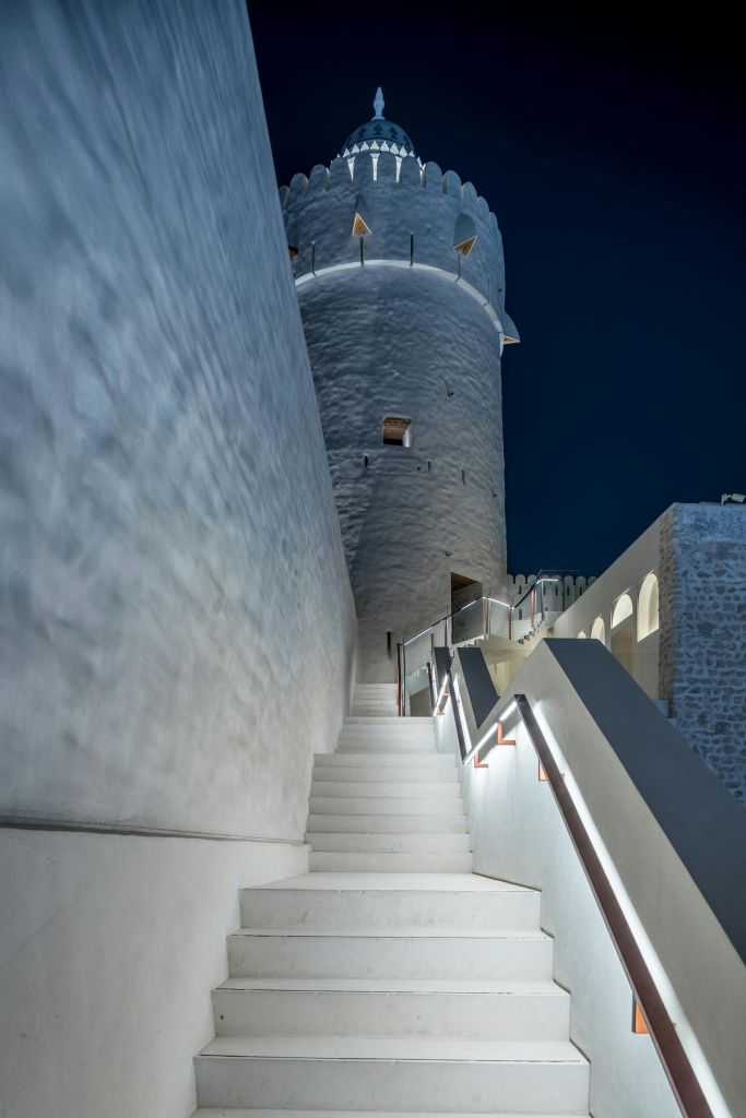 조명이 켜진 계단을 따라 어두운 밤하늘 아래 낡고 하얀 돌탑으로 이어집니다. 벽과 난간에는 부드러운 조명이 켜져 있습니다.