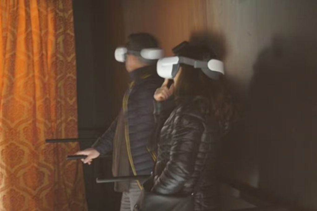 조명이 어두운 방에서 가상 현실 헤드셋을 착용한 두 사람이 화려한 커튼이 보이는 벽 근처에 서 있습니다.