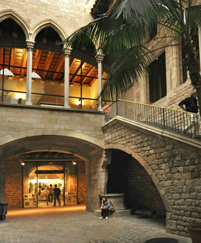 위층에 석조 아치, 계단, 갤러리가 있는 실내 안뜰. 아래 벤치에 두 사람이 앉아 있고, 안쪽에는 다른 사람들이 보입니다.