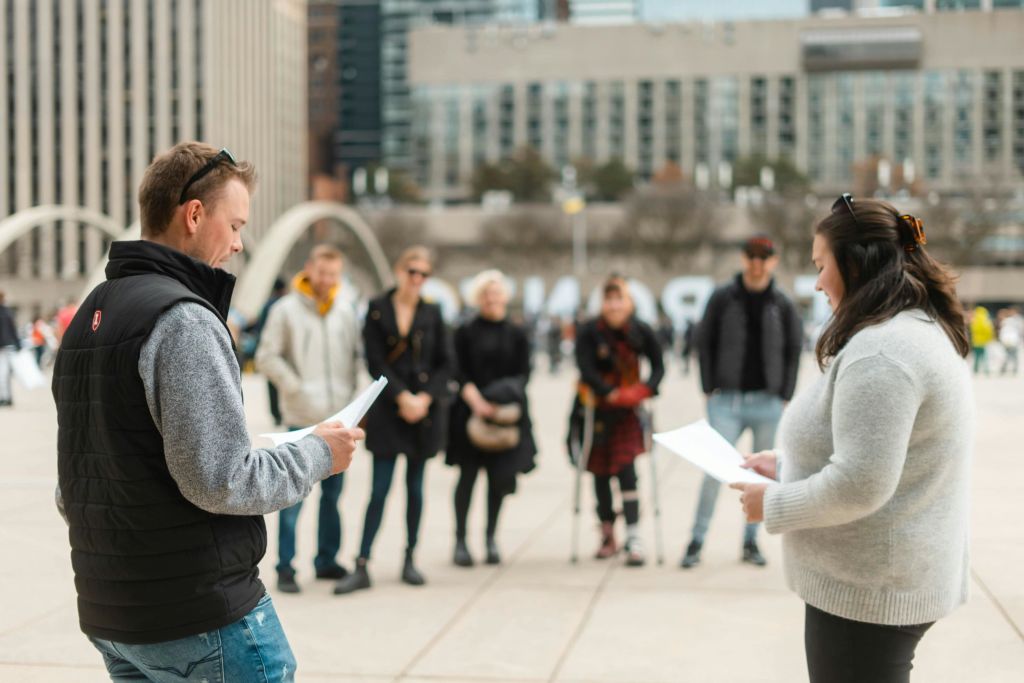 한 남성이 야외 도심 광장의 종이를 읽고 있는데, 배경에는 여러 사람이 서 있습니다.