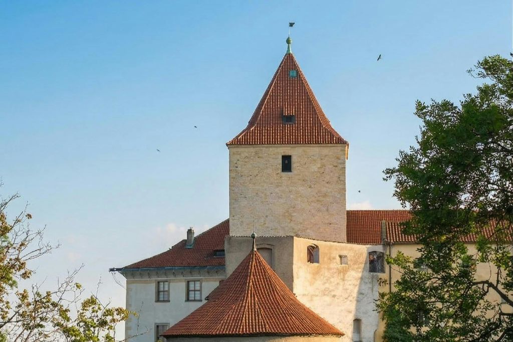 붉은 기와 지붕이 있는 유서 깊은 석조 건물과 나무로 둘러싸인 맑고 푸른 하늘 아래 눈에 띄는 탑이 있습니다.