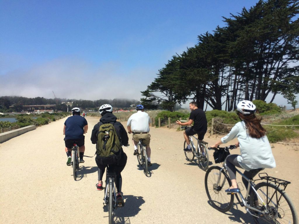 화창하고 부분적으로 흐린 하늘 아래 녹음과 나무로 둘러싸인 자갈길에서 자전거를 타는 사람들의 모임.