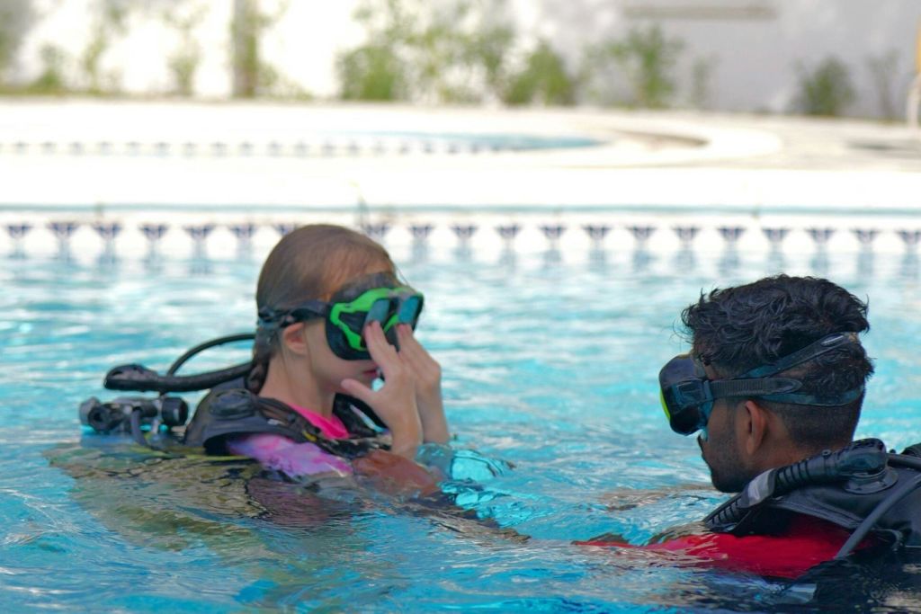 스쿠버 장비를 착용한 두 사람이 수영장에 있으며, 한 명은 마스크를 조정하고 다른 한 명은 마스크를 착용하고 있습니다.