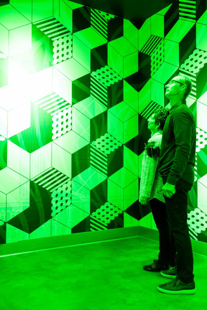 두 사람이 녹색 기하학적 무늬의 벽 앞에 서서 밝은 빛을 바라보고 있습니다.