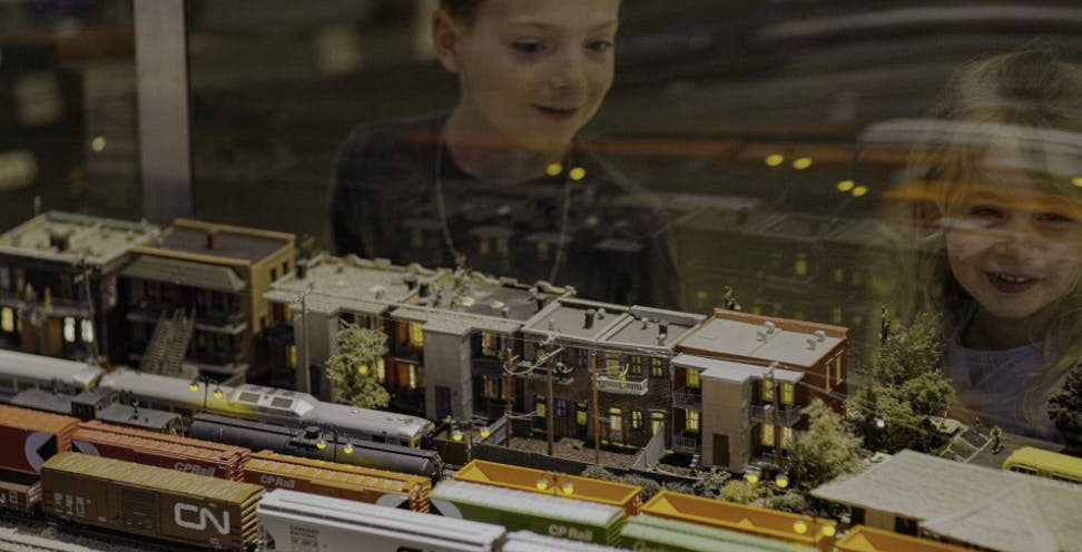 두 어린이는 유리 인클로저를 통해 건물과 녹지가 있는 상세한 모형 열차 디스플레이를 흥미롭게 바라보고 있습니다.