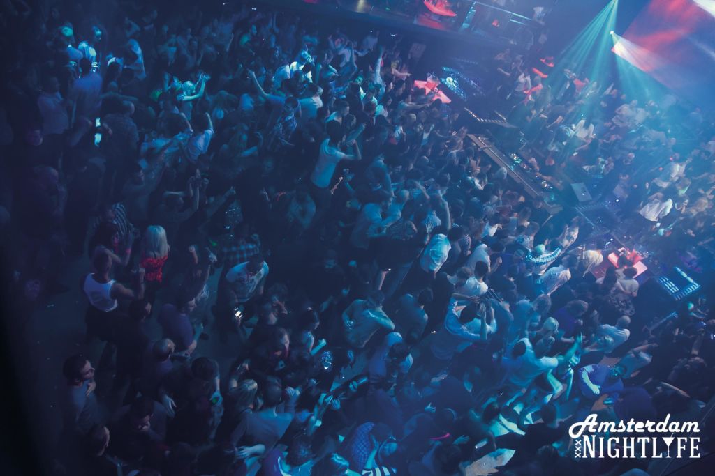 파란색과 보라색 조명 아래에서 춤을 추는 사람들이 있는 붐비는 나이트클럽 댄스 플로어와 DJ 부스가 배경으로 보입니다.