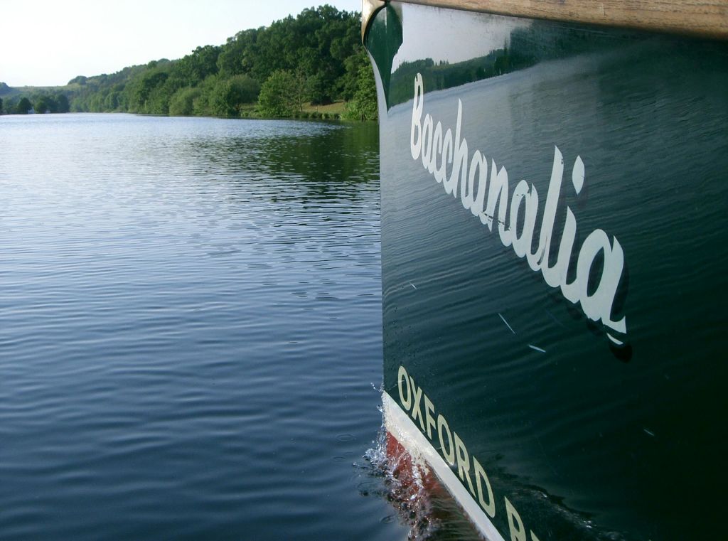 "옥스포드" 가 보이는 "바카날리아" 라는 이름의 녹색 보트의 측면은 숲이 우거진 해안가 근처의 잔잔한 물을 통과합니다.