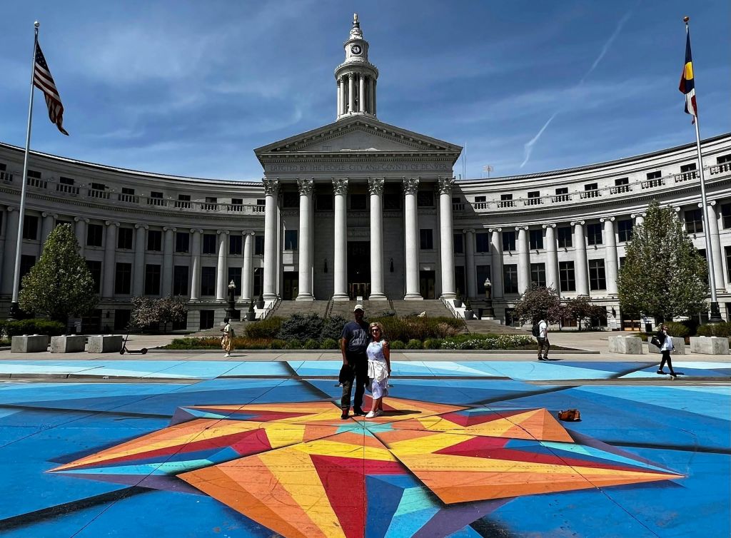 두 사람이 커다란 신고전주의 양식의 건물을 배경으로 광장에서 화려한 별 모양의 예술 작품 위에 서 있습니다.