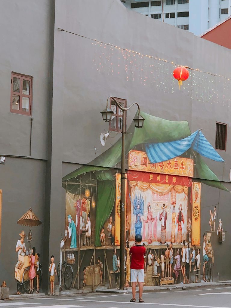 의상을 입은 배우들과 전통 공연을 묘사한 벽 벽화로, 빨간 등불이 머리 위로 놓인 채 관람하는 사람들로 둘러싸여 있습니다.