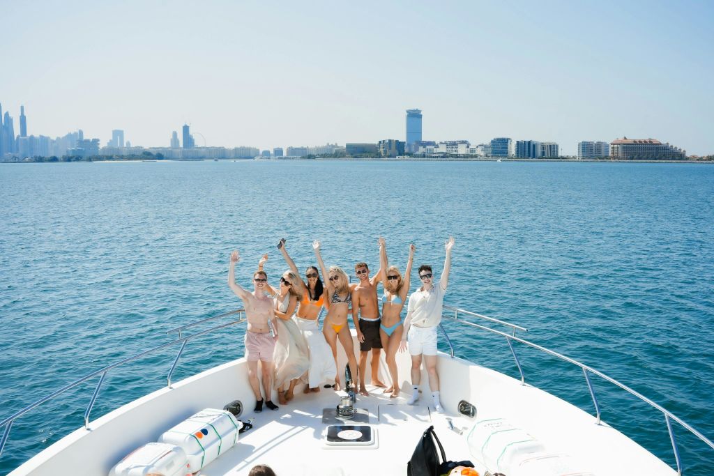 수영복을 입은 7명이 보트 갑판에 서서 도시 풍경과 푸른 물을 배경으로 포즈를 취하고 손을 흔들고 있습니다.