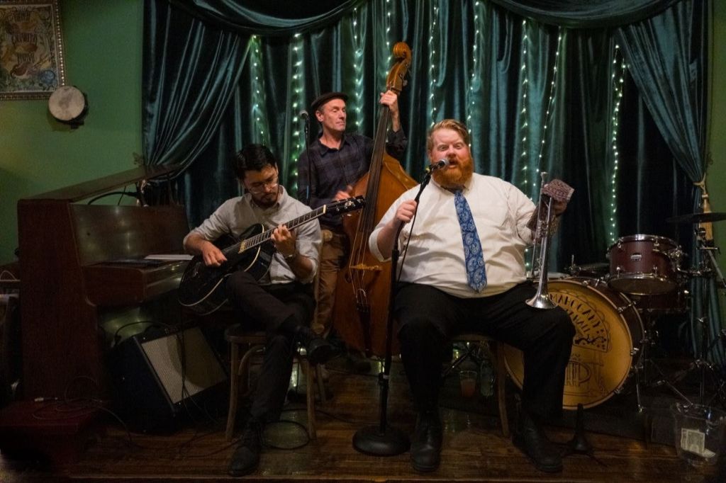 세 명의 뮤지션이 무대에서 공연합니다. 한 명은 기타를 연주하고, 한 명은 직립 베이스를 연주하고, 한 명은 트럼펫을 들고 마이크를 불렀습니다.