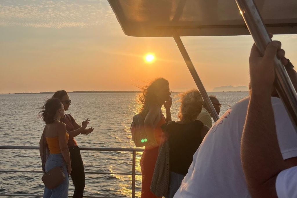 해가 질 무렵 보트 갑판에 사람들이 서 있는데, 해는 하늘에서 낮고 바다를 배경으로 잔잔한 바다가 펼쳐집니다.