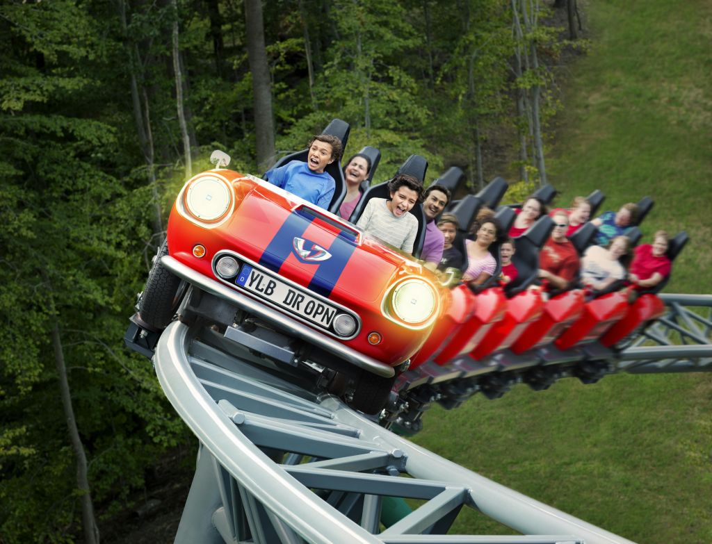 나무와 잔디로 둘러싸인 미니 쿠퍼 모양의 빨간색과 파란색 롤러코스터 자동차를 타고 스릴 넘치는 라이딩을 즐기는 사람들.