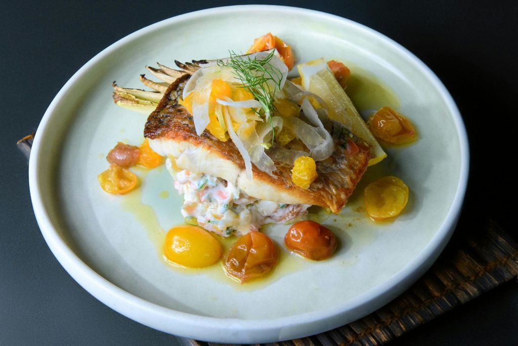 토핑, 체리 토마토, 레몬 웨지가 담긴 생선 구이가 담긴 도금 요리로, 밝은 색상의 접시에 담겨 나옵니다.