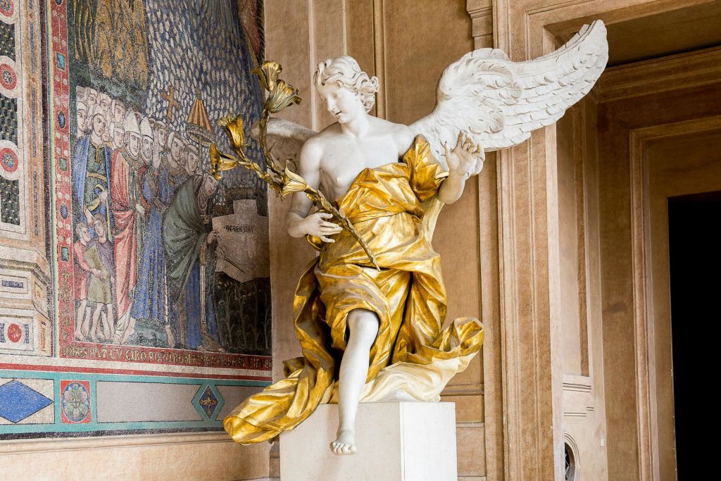 웅장한 건물 내부의 복잡한 모자이크 벽에 세워진 백합을 들고 있는 금색 천으로 장식된 우아한 날개 동상.