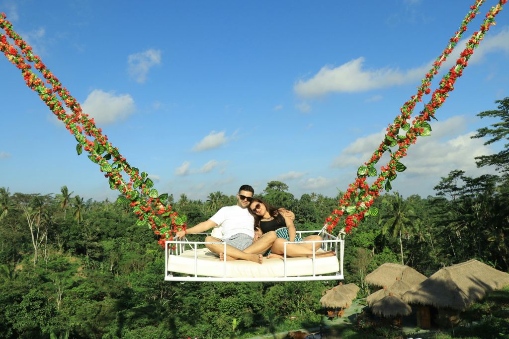 맑고 푸른 하늘과 구름을 배경으로 울창한 녹음 위에 매달려 있는 꽃 장식 그네에 한 커플이 앉아 있습니다.