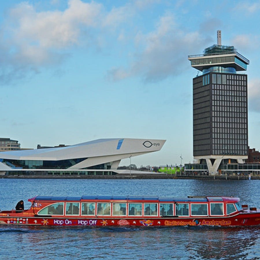 푸른 하늘 아래 현대적인 건물과 높은 탑을 배경으로 강에서 "홉 온 홉 오프 (Hop On Hop Off)" 라는 이름의 빨간색 투어 보트가 있습니다.