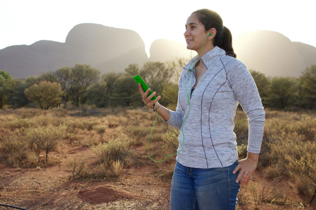 야외에서 이어버드를 착용하고, 스마트폰을 들고, 미소를 짓고, 사막 풍경과 멀리 떨어진 암석을 배경으로 활동하는 사람.