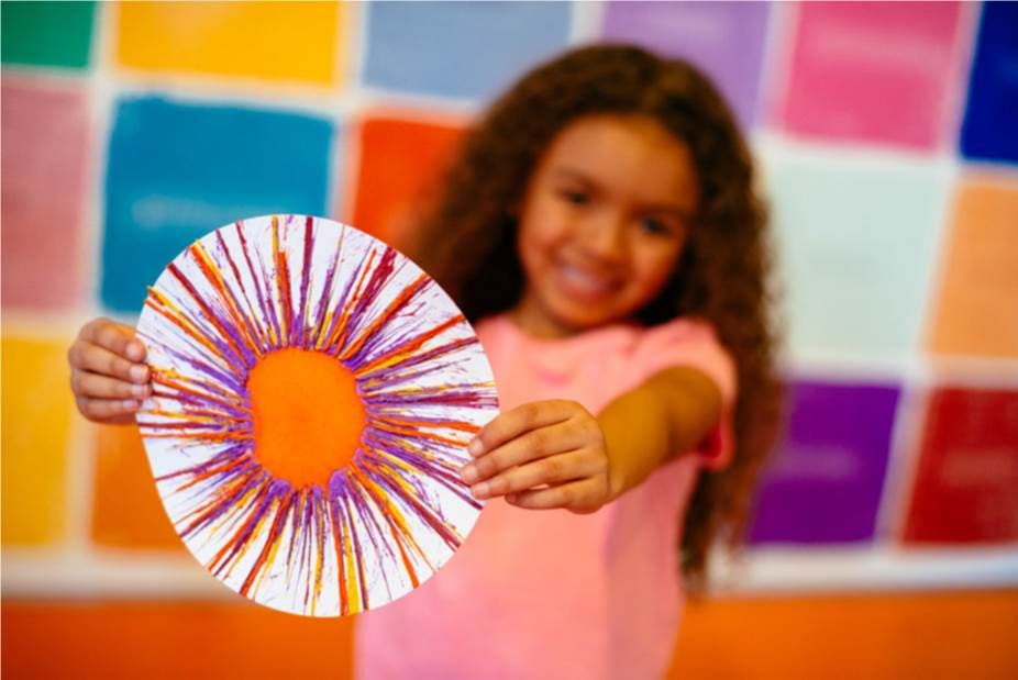 밝은 색상의 배경 앞에서 화려한 휠 모양의 그림을 들고 웃으며 분홍색 셔츠를 입은 아이.