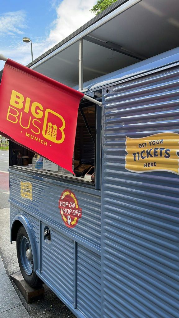 파란색 차량의 티켓 부스에는 'Big Bus Munich', 'Get Your Tickets Here', 'Hop On Hop Off' 표지판이 있습니다.