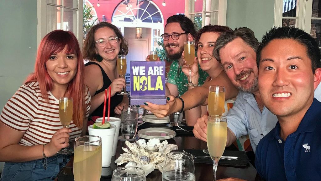 한 무리의 사람들이 음료와 함께 테이블에 앉아 "We Are NOLA" 라고 적힌 팻말을 들고 있습니다.