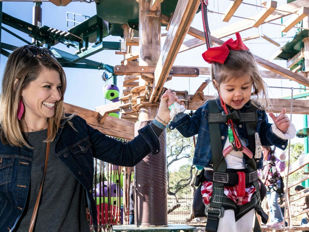 하네스의 어린 소녀가 로프 코스를 탐색하는 동안 어른의 손을 잡고 웃으며 빨간색 활을 착용합니다.