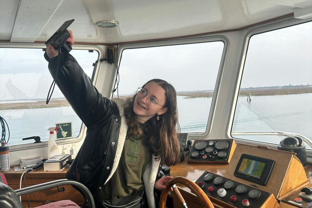 배의 조종석에 서 있는 사람이 셀카를 찍습니다. 배경은 커다란 창문을 통해 물과 육지를 보여줍니다.