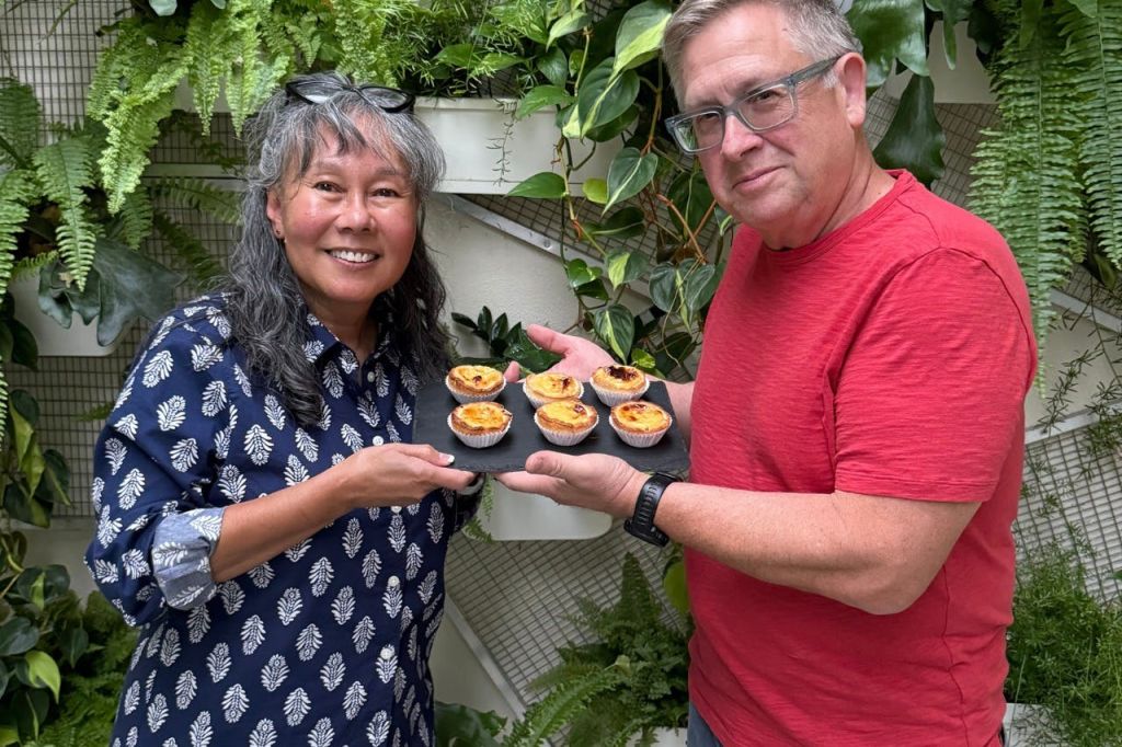 녹음이 우거진 식물을 배경으로 페이스트리가 담긴 트레이를 들고 웃고 있는 두 사람.