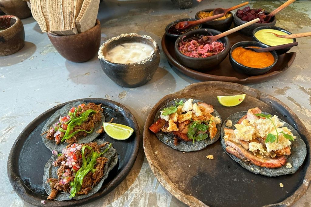 다양한 토핑과 라임 웨지가 담긴 타코 두 접시, 화이트 음료 한 그릇, 냅킨, 사이드와 소스 그릇.