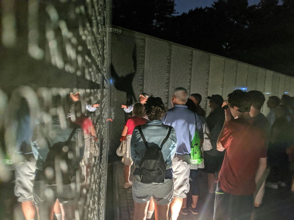 야간에 조명이 켜진 이름들이 새겨진 반사 벽 주위에 사람들이 모여듭니다. 가이드가 비문을 향해 제스처를 취합니다.