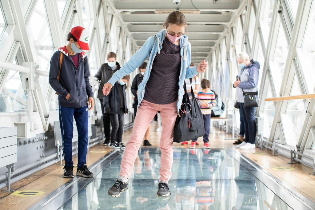 마스크를 착용한 사람들이 실내에서 유리 바닥 다리를 걷고 있습니다. 한 여성이 중앙에서 조심스럽게 균형을 잡습니다.