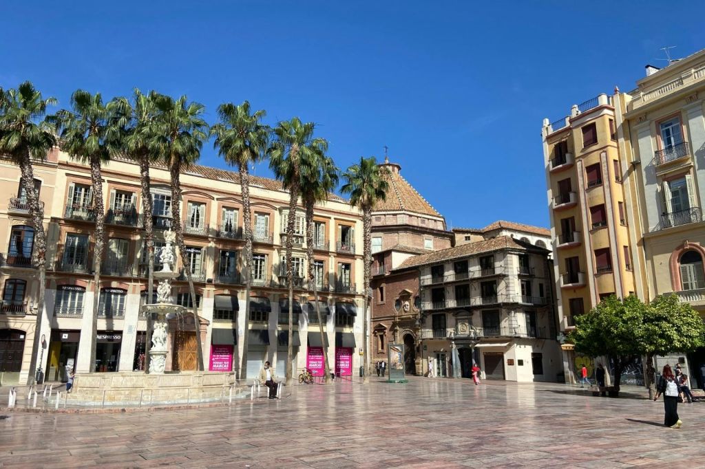 야자수, 유서 깊은 건물, 분수, 보행자가 거의 없는 햇살 가득한 타운 광장. 푸른 하늘을 배경으로.