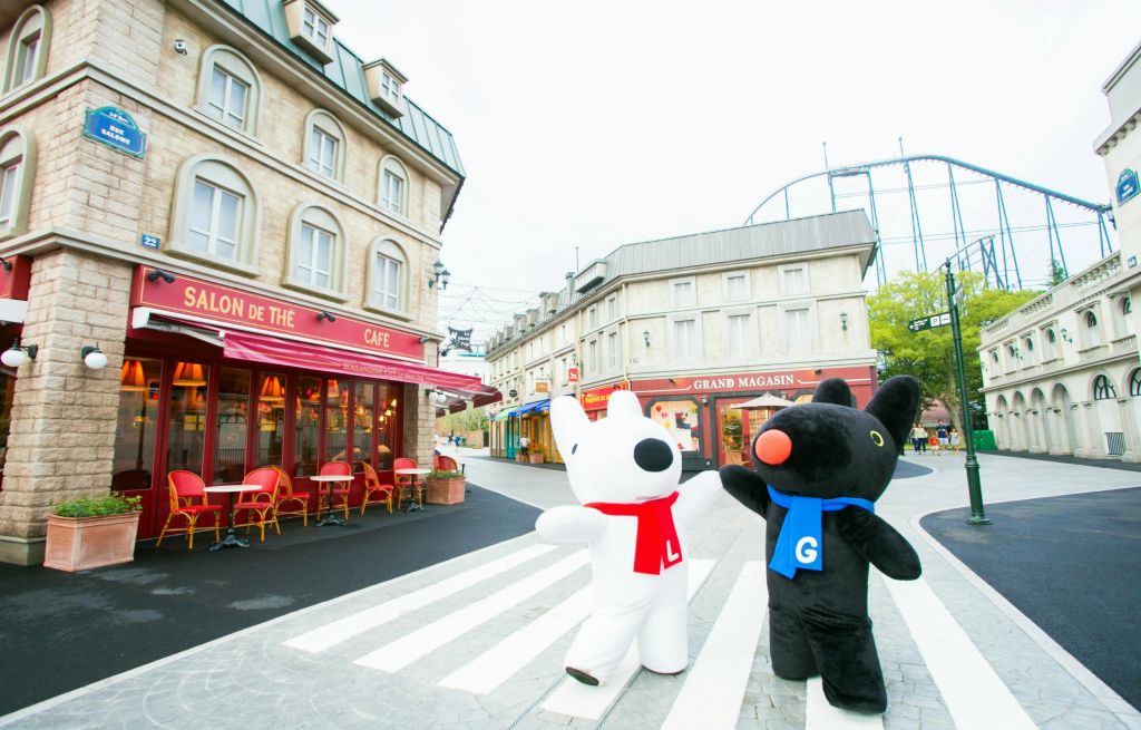 두 명의 마스코트 캐릭터가 빨간색 스카프를 두른 흰색 스카프와 파란색 스카프를 두른 검은색 스카프를 두른 검은색 스카프가 독특한 거리 풍경의 횡단보도에 서 있습니다.