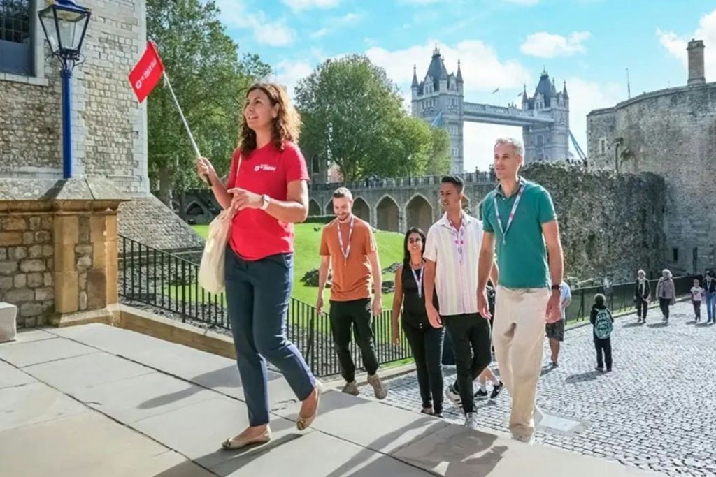 투어 가이드가 일행과 함께 런던 타워를 지나갑니다. 화창한 날에는 타워 브리지가 배경으로 보입니다.