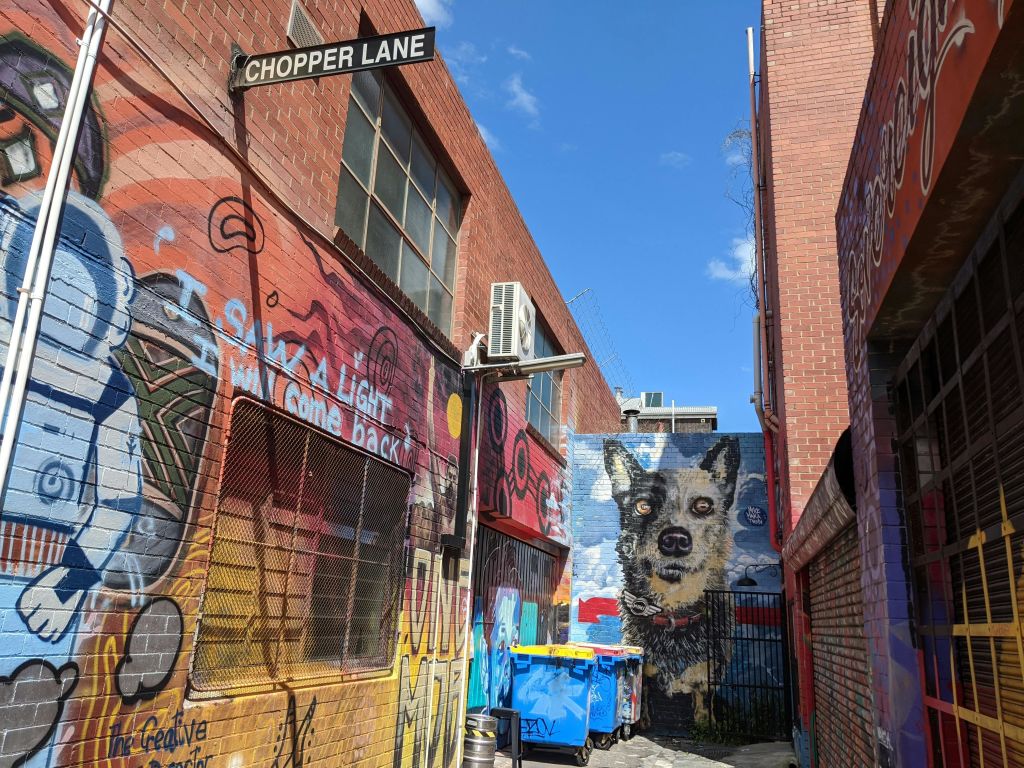 알록달록한 낙서, 대형 개 벽화, 벽돌 건물, 에어컨, 쓰레기통, "Chopper Lane" 표지판이 있는 좁은 골목.