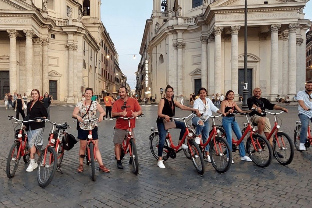자갈길에 기둥이 있는 유서 깊은 건물 앞에서 6명의 일행이 빨간 자전거를 타고 있습니다.