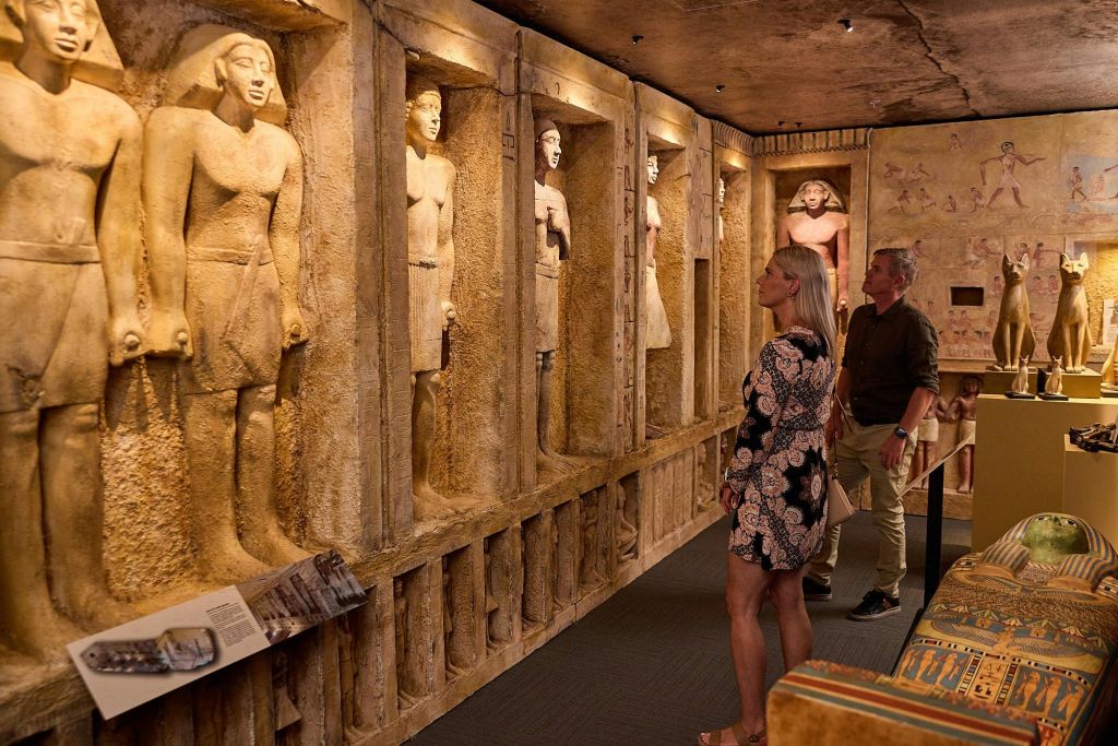 박물관 전시관의 벽 틈새에 있는 고대 이집트 동상. 한 사람이 오른쪽 디스플레이를 관찰하고 있습니다.